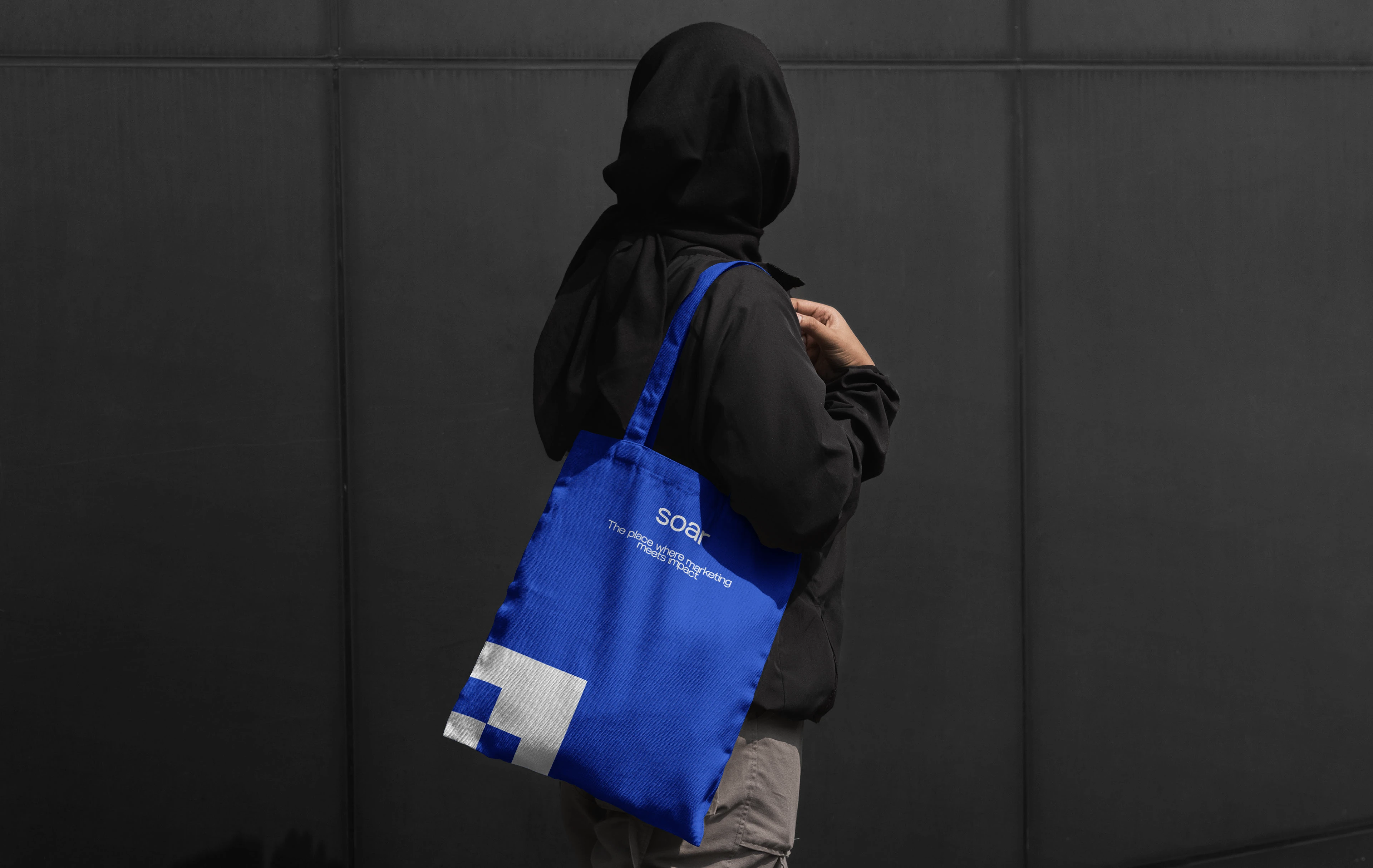 Soar blue tote bag carried by a person in a dark hooded jacket